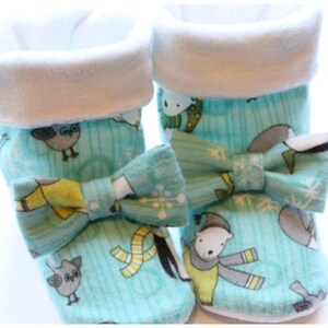 May include: A pair of baby booties with a winter-themed pattern. The booties are light blue with illustrations of owls, polar bears, and snowflakes. Each bootie has a white cuff and a matching bow. The booties are made of soft fabric.