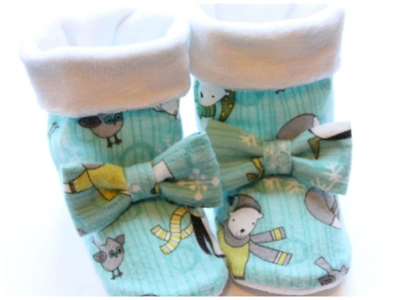 May include: A pair of baby booties with a winter-themed pattern. The booties are light blue with illustrations of owls, polar bears, and snowflakes. Each bootie has a white cuff and a matching bow. The booties are made of soft fabric.