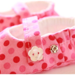 May include: Pink baby shoes with a polka dot pattern in shades of pink and red. The shoes have a white interior and a strap with a flower-shaped button closure. The shoes are designed for infants.