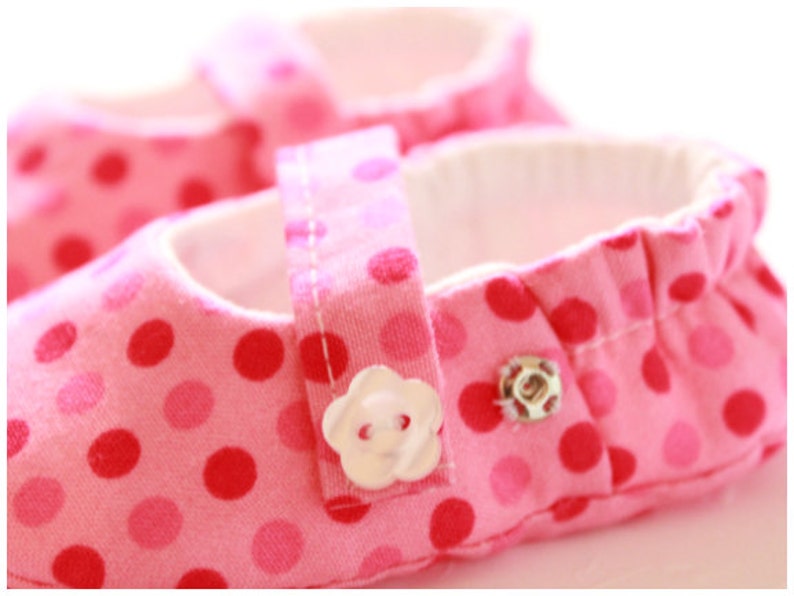 May include: Pink baby shoes with a polka dot pattern in shades of pink and red. The shoes have a white interior and a strap with a flower-shaped button closure. The shoes are designed for infants.