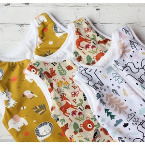May include: Four baby sleep sacks with various patterns. One is mustard yellow with animal illustrations, another has a woodland theme with deer and bears, and the third features unicorns and trees. Each sack has a white interior and a zipper closure.