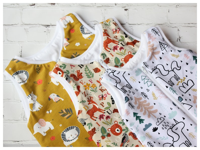 May include: Four baby sleep sacks with various patterns. One is mustard yellow with animal illustrations, another has a woodland theme with deer and bears, and the third features unicorns and trees. Each sack has a white interior and a zipper closure.