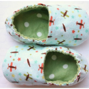 May include: A pair of baby booties with a light blue exterior patterned with brown and red airplanes, yellow stars, and green stars. The interior is green with white polka dots, and the trim is a darker green.