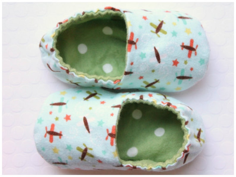 May include: A pair of baby booties with a light blue exterior patterned with brown and red airplanes, yellow stars, and green stars. The interior is green with white polka dots, and the trim is a darker green.