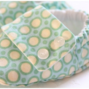 May include: Close-up of a baby shoe with a light blue fabric patterned with green and cream-colored circles. The shoe has a flap secured with two small white buttons. The interior is white.
