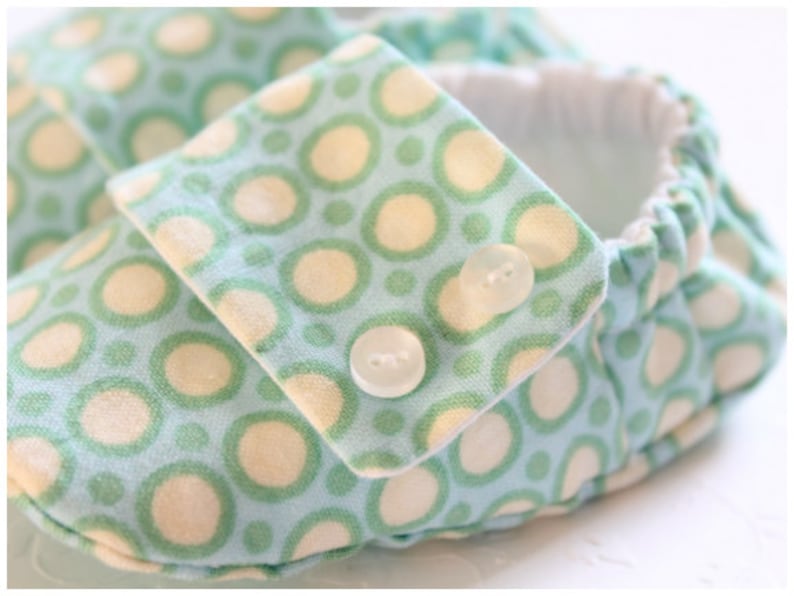 May include: Close-up of a baby shoe with a light blue fabric patterned with green and cream-colored circles. The shoe has a flap secured with two small white buttons. The interior is white.