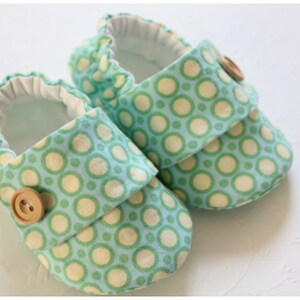 May include: A pair of baby shoes with a light blue background and a pattern of green and cream-colored circles. Each shoe has a flap secured by a wooden button. The interior is white.