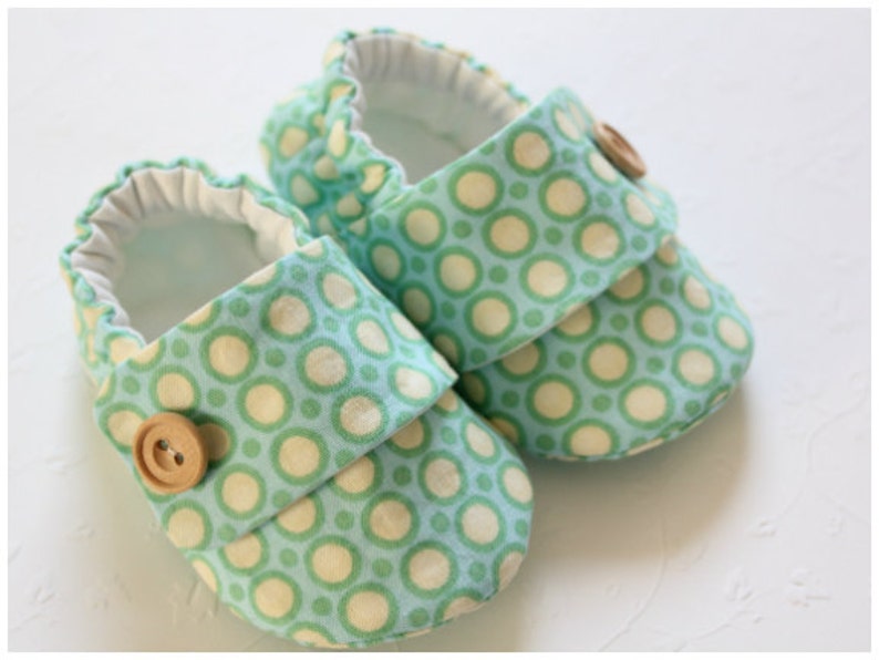 May include: A pair of baby shoes with a light blue background and a pattern of green and cream-colored circles. Each shoe has a flap secured by a wooden button. The interior is white.