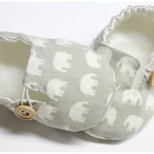 May include: A pair of baby shoes featuring a grey elephant pattern. The shoes have a soft, white interior and are fastened with a wooden button and elastic loop. These shoes are designed for comfort and style.