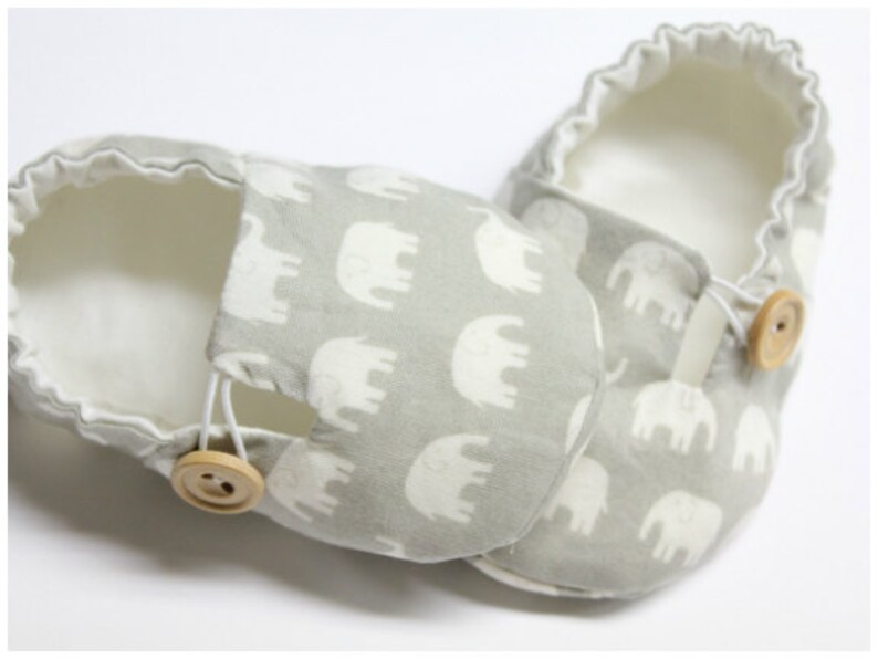 May include: A pair of baby shoes featuring a grey elephant pattern. The shoes have a soft, white interior and are fastened with a wooden button and elastic loop. These shoes are designed for comfort and style.