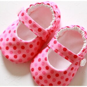 May include: A pair of pink baby shoes with a polka dot pattern. The shoes have a strap with a button closure and a ruffled trim around the opening. The interior is white.