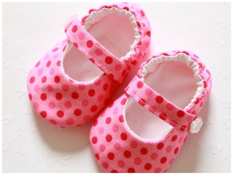 May include: A pair of pink baby shoes with a polka dot pattern. The shoes have a strap with a button closure and a ruffled trim around the opening. The interior is white.