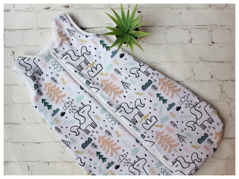 May include: White baby sleep sack with a whimsical unicorn and forest pattern. The design includes outlines of unicorns, trees, clouds, and geometric shapes in black, peach, green, and blue. A white zipper runs down the center.