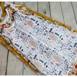 May include: Several baby sleep sacks with different patterns are stacked on top of each other. The top sleep sack has a white background with a unicorn and forest design. Other sleep sacks have animal and floral patterns. The sleep sacks have a zipper closure.
