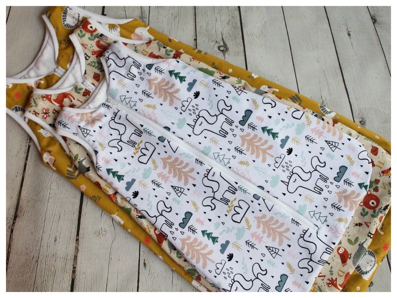 May include: Several baby sleep sacks with different patterns are stacked on top of each other. The top sleep sack has a white background with a unicorn and forest design. Other sleep sacks have animal and floral patterns. The sleep sacks have a zipper closure.