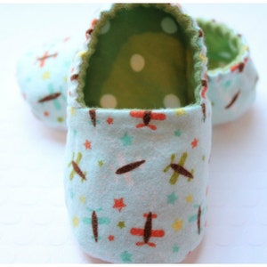 May include: Baby booties in a light blue fabric with a repeating pattern of airplanes, stars, and dots. The interior is green with white polka dots. The edges are trimmed with a contrasting stitch.