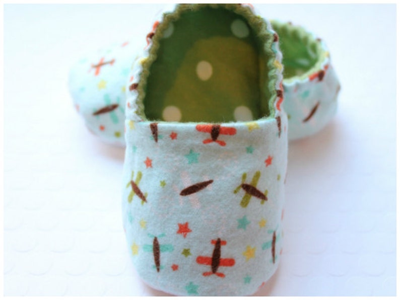 May include: Baby booties in a light blue fabric with a repeating pattern of airplanes, stars, and dots. The interior is green with white polka dots. The edges are trimmed with a contrasting stitch.