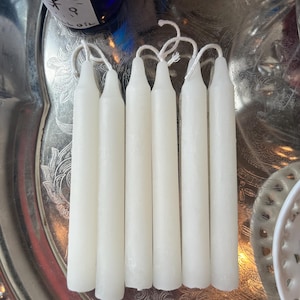 May include: Six white taper candles arranged in a vertical row. Each candle features a smooth, cylindrical body that tapers slightly toward the top, ending in a pointed tip with a short, white cotton wick extending from the center of the top.