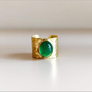 Op de afbeelding: Een brede, goudkleurige metalen ring met een gehamerd, getextureerd oppervlak en een open ontwerp aan de achterkant. Het midden van de ring is bezet met een gladde, ovaalvormige, levendig groene cabochonsteen met een hoogglans afwerking.