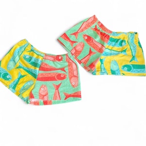 May include: Two pairs of shorts featuring a repeating fish pattern. One pair has a yellow background with teal fish, while the other has a light green background with red fish. Both garments have an elasticized waistband and a simple hemmed leg opening.