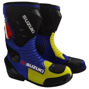 Puede incluir: Un par de botas de carreras para motocicleta en azul, amarillo y negro. Las botas presentan el nombre de la marca Suzuki en texto blanco en el lateral y la espinillera, con un acento de logotipo rojo. El diseño incluye paneles protectores negros.