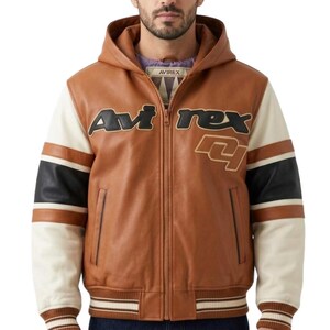 Puede incluir: Una chaqueta de cuero beige con capucha que presenta el texto Avirex en letras negras en el pecho y un gran gráfico delineado debajo. Las mangas tienen bloques de color con rayas crema, negras y beige, y el dobladillo y los puños son acanalados.