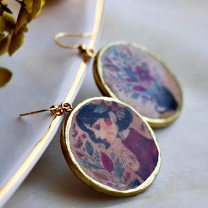 May include: A pair of round drop earrings featuring gold-toned metal frames. Each earring displays a printed illustration of a person with dark hair surrounded by colourful floral and botanical elements in shades of red, purple, and green.