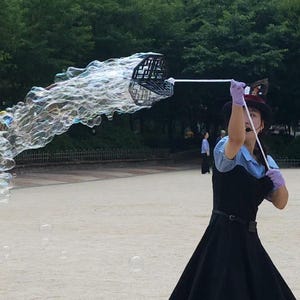 May include: A performer wearing a black dress with a light blue shirt underneath, a black hat, and light purple gloves, holding a black bubble wand that is creating a large stream of translucent soap bubbles in an outdoor setting.