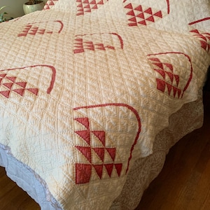 May include: A cream-colored quilt featuring a repeating pattern of red basket motifs. The fabric has a diamond-shaped quilted texture throughout, with the basket designs appliqued in red fabric across the surface of the bed covering.