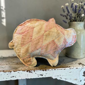 May include: A stuffed pig toy crafted from quilted fabric in a diamond pattern. The textile features a faded color palette of soft peach, cream, and pale pink tones with visible stitching throughout the surface, creating a textured appearance.