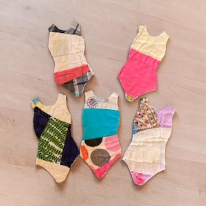May include: Five fabric cutouts shaped like one-piece swimsuits. One features a plaid pattern, one is solid pink and cream, one has green and navy geometric shapes, one displays teal and polka dots, and one shows a floral print with cream fabric.