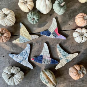 May include: Five quilted fabric witch hat ornaments in various colors and patterns, including blue, yellow, pink, and plaid, arranged among several small decorative pumpkins of different colors and textures on a wooden surface.