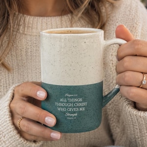 May include: A two-toned ceramic mug with a speckled cream top and a dark teal bottom. The teal section features the white text: Philippians 4:13 ALL THINGS THROUGH CHRIST WHO GIVES ME Strength Philippians 4:13. The mug has a curved handle.