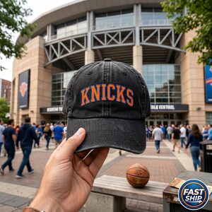 May include: A charcoal grey baseball cap with a distressed, washed-out finish. The word KNICKS is embroidered across the front in bold orange letters with a blue outline. The cap features a curved brim and a structured six-panel crown design.