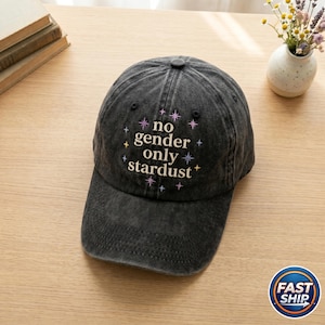 May include: A charcoal grey baseball cap with a distressed finish featuring the embroidered text 'no gender only stardust' in a light cream color, surrounded by small, multi-colored star icons in shades of purple, yellow, and light blue.