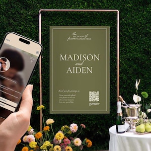 Op de afbeelding: Een smartphone met een digitale uitnodigingsapp naast een verticaal olijfgroen bord met de tekst "The beginning of forever starts here," "MADISON and AIDEN," en een QR-code, gemonteerd op een koperen standaard.