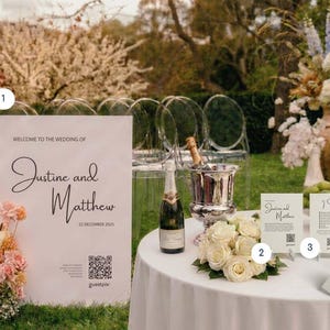 Könnte beinhalten: Ein weißes Hochzeitsschild mit schwarzem Text Justine and Matthew 22 December 2025 und QR-Code, ein runder Tisch mit weißer Tischdecke, eine Champagnerflasche, ein silberner Sektkühler, weiße Rosen und drei kleine Informationskarten.