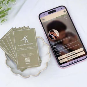 Könnte beinhalten: Ein Stapel rechteckiger olivgrüner Karten mit weißem Text M&A Madison & Aiden und einem QR-Code, neben einem Smartphone, das eine digitale Einladung mit einem unscharfen Foto eines Paares und Schaltflächen zum Hochladen von Fotos anzeigt.