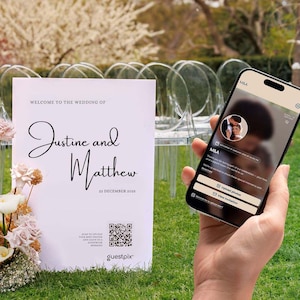 Könnte beinhalten: Ein weißes Hochzeitsschild mit schwarzem Text Welcome to the wedding of Justine and Matthew 22 December 2023 und einem QR-Code, neben einem Smartphone, das eine digitale Veranstaltungsoberfläche mit einem runden Foto und Schaltflächen zeigt.