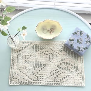 May include: A rectangular cream-colored crochet doily with an openwork pattern, a small white ceramic dish containing two metal rings, a light blue square box with white floral embroidery, and a clear glass vase with white artificial flowers.