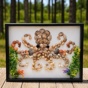 May include: A framed wall art piece featuring an octopus mosaic crafted from various small brown, tan, and grey seashells on a white background. Two small green artificial plants are positioned at the bottom corners of the black rectangular frame.