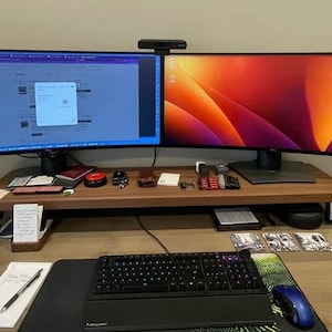 May include: A modern home office workspace with two curved monitors, a keyboard, and a mouse. The monitors are on a wooden stand and the desk is made of wood. There are various items on the desk, including a red notebook, a black wallet, and a pair of headphones.