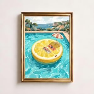 May include: A framed art print depicting a large yellow lemon slice pool float in turquoise water. Two figures lie on towels on the float under a striped umbrella. The background shows a coastal landscape with trees, hills, and a blue sea.