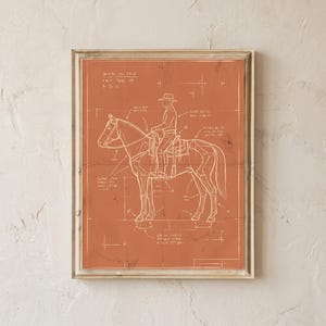 May include: A framed wall art print featuring a terracotta-colored background with a white line drawing of a cowboy riding a horse. The illustration includes anatomical skeletal details, technical drafting lines, and handwritten notes in white ink.