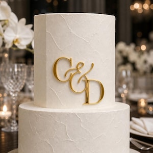 May include: A two-tiered white cake with a textured, matte finish. The top tier is hexagonal, while the bottom tier is cylindrical. A shiny, gold-coloured acrylic monogram featuring the letters C and D joined by an ampersand is attached to the front.