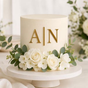 May include: A cylindrical cake with smooth off-white frosting, featuring gold-colored capital letters A and N separated by a vertical line. The base is decorated with a cluster of white roses, small white flowers, and green eucalyptus leaves.
