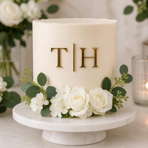 Op de afbeelding: Een cilindrische taart met glad, gebroken wit glazuur, voorzien van goudkleurige letters T en H gescheiden door een verticale lijn. De basis is versierd met een boeket witte rozen, kleine witte bloemen en groene eucalyptusbladeren op een marmeren voet.