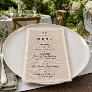 May include: A rectangular beige fabric menu card with frayed edges, featuring black printed text that reads MQ MENU, FIRST COURSE Smoked Beet Salad, MAIN COURSE Rosemary Bison Short Rib, DESSERT Maple Bourbon Bread Pudding, MAEVE & QUINN, NOVEMBER 26, 2026.