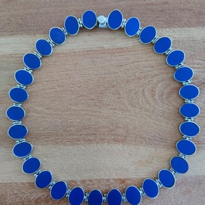 Op de afbeelding: Een cirkelvormige ketting bestaande uit achtentwintig ovale schakels, elk met een gladde, effen blauwe inleg in een zilverkleurig metalen frame. De schakels zijn verbonden door kleine scharnieren en het sieraad is voorzien van een metalen sluiting.