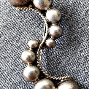 Könnte beinhalten: Eine silberfarbene Brosche mit einem geschwungenen S-förmigen Design, das aus mehreren glatten, kugelförmigen Perlen unterschiedlicher Größe besteht. Ein gedrehter Metalldraht verläuft entlang der Innenkurve der Perlen und verbindet sie.